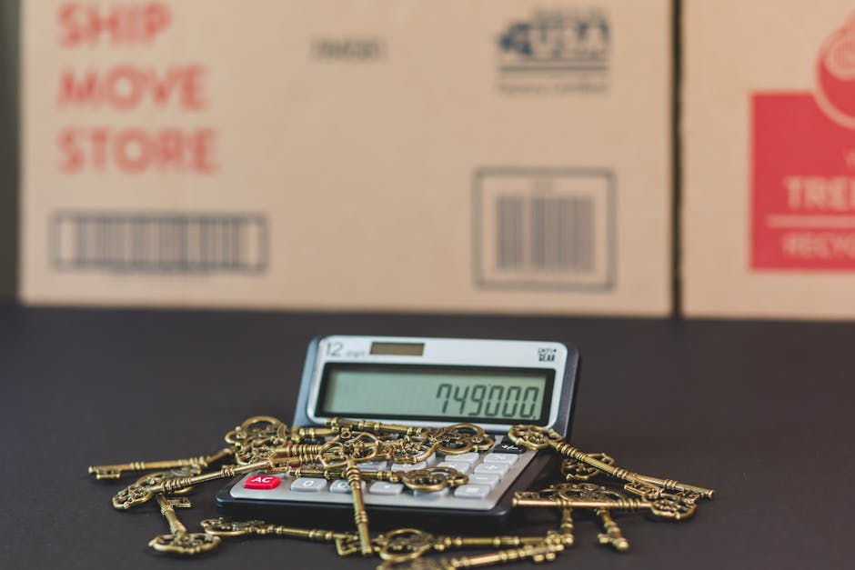 A close-up image showing a black calculator with a large digital display reading '749800', surrounded by multiple gold-colored keys on a dark surface. In the background, there are cardboard boxes with printed labels and barcodes, indicating a moving or packing environment. The boxes are stacked inside a property near a doorway, suggesting a home relocation process. The setup illustrates the packing and moving phase typical of house removals, with a focus on attention to detail in handling valuables and organized logistics, as handled by Removal Company Highbury. The scene emphasizes the importance of secure packing materials and proper documentation during furniture transport and home moving services.