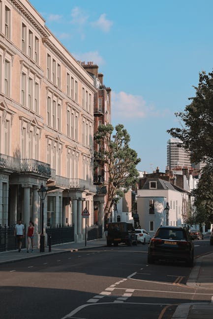 A street scene showing a row of multi-storey residential buildings with classic architectural details, large windows, and balconies, situated along a paved sidewalk with pedestrians walking nearby. Several parked cars are visible along the curb, and a black van is parked close to the buildings, potentially used for furniture transport. In the foreground, a small collection of cardboard boxes, wrapped furniture, and plastic-clarified items are arranged on the pavement as part of a home relocation process. Additionally, some furniture items, such as a wooden wardrobe and a cardboard box filled with packing materials, are being carried or prepared for loading onto a van operated by Removal Company Highbury, with the interior of the property partially visible through an open doorway. The scene is set during daylight with a clear blue sky, and trees are present along the street, reflecting the urban environment ready for a professional moving or packing service.