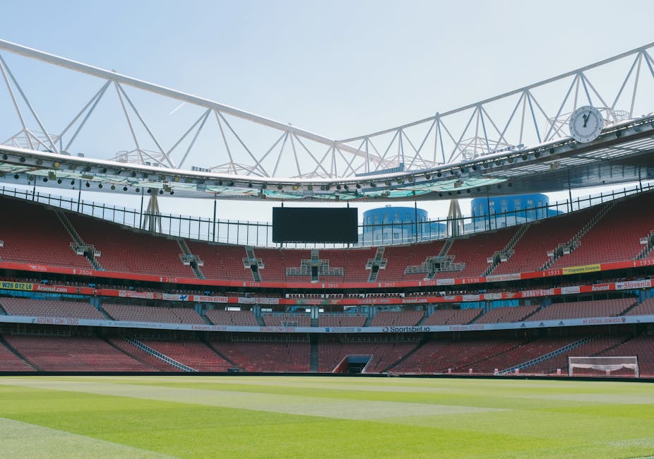 The image shows the interior of a modern football stadium with a large, well-maintained grass pitch and surrounding tiered seating in red and grey, designed for spectators. The open roof structure is supported by white steel trusses, allowing natural light to illuminate the field. Visible features include a large central digital scoreboard above the seats, empty spectator stands, and some advertising banners along the perimeter. The stadium is clean and appears to be prepared for a match or event. This setting may relate to the logistics of sports-related moves or equipment transport, aligning with commercial removals and furniture transport services provided by Removal Company Highbury, particularly for relocation or fixture installation at sports venues. The overall scene emphasizes the scale of the venue and the structural elements involved in supporting large-scale sporting events, with an emphasis on unobstructed views and accessibility considerations.