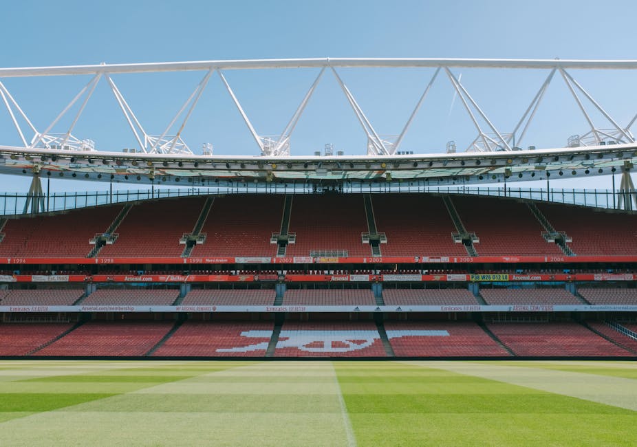 Image showing an empty section of a sports stadium with red seating and a large, white, modern canopy structure overhead. A round clock is mounted on a supporting beam of the canopy, displaying approximately 2:55. There are metal railings and some dark electronic screens, possibly for display purposes. The scene suggests a preparation or maintenance activity related to stadium operations, possibly during a move or setup process for a sporting event. This setting is outside a stadia environment, consistent with the logistics involved in house or commercial relocations, such as those managed by Removal Company Highbury, which provides professional move and transport services. The image emphasizes the structural aspects, seating arrangement, and the open-air nature of the venue, with daylight illumination and a clear sky in the background.