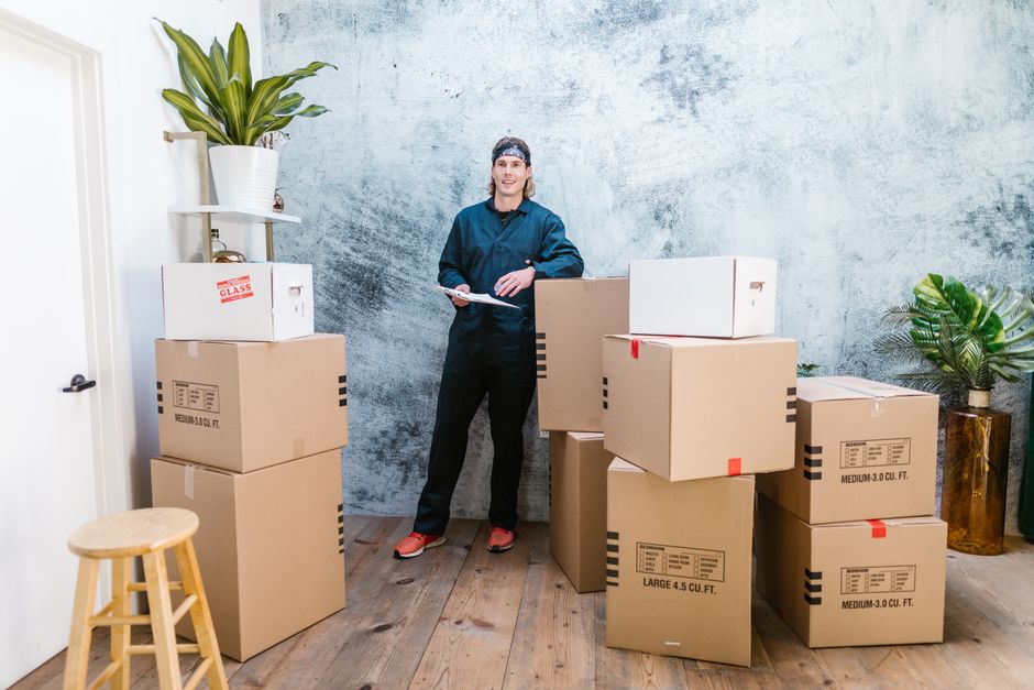Two individuals are sitting on a hardwood floor during a home relocation process, with a collection of packed books both inside and outside a cardboard box labeled 'BOOKS' with a marker. One person is