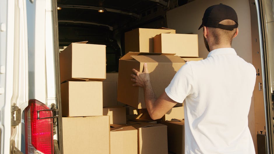 A man wearing a white polo shirt and a black cap is standing inside the open rear of a white van, which is parked on a paved area. He is in the process of loading or unloading multiple cardboard boxes of various sizes, some of which are partially wrapped in plastic or packing paper, onto or from the vehicle. The boxes are arranged in a stacked manner, with the man holding a medium-sized box close to his chest, ready to place it inside the van or lift it out. The interior of the van is dimly lit, but the outgoing daylight illuminates the scene, highlighting the boxes and the man's actions related to home relocation or furniture transport. Removal Company Highbury offers professional removals and moving services, including packing and loading processes, to facilitate efficient house moves in the Highbury area.