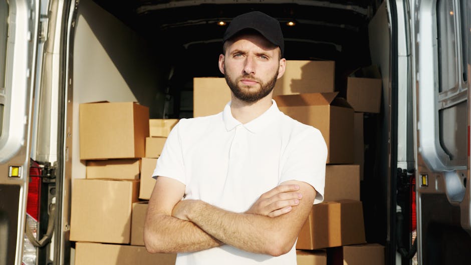 A man with a neatly trimmed beard, wearing a white polo shirt and a black cap, stands with his arms crossed inside the open back of a moving van. The van is filled with stacked cardboard boxes of various sizes, some wrapped in plastic or packing paper, ready for a house removal or relocation. The interior of the van is well-lit, showing the wooden floor and structural elements, with the boxes arranged in an orderly manner to facilitate efficient loading and transport. Outside the van, the pavement and part of the exterior vehicle body are visible, indicating the scene takes place during a home relocation process. This setup highlights the furniture transport and packing stage involved in professional removals, as provided by Removal Company Highbury, aligning with house removals and moving logistics themes.