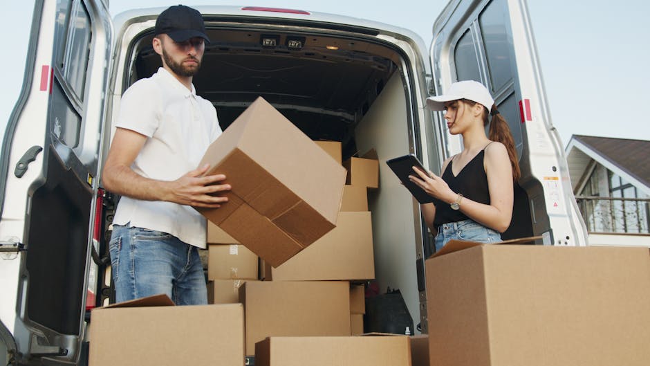 A man wearing a white polo shirt and a black cap is standing inside the open rear of a white van, which is parked on a paved area. He is in the process of loading or unloading multiple cardboard boxes of various sizes, some of which are partially wrapped in plastic or packing paper, onto or from the vehicle. The boxes are arranged in a stacked manner, with the man holding a medium-sized box close to his chest, ready to place it inside the van or lift it out. The interior of the van is dimly lit, but the outgoing daylight illuminates the scene, highlighting the boxes and the man's actions related to home relocation or furniture transport. Removal Company Highbury offers professional removals and moving services, including packing and loading processes, to facilitate efficient house moves in the Highbury area.
