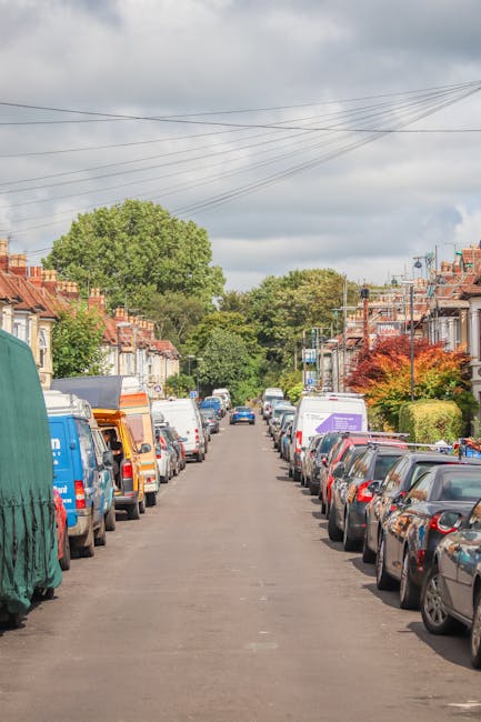 A quiet residential street in Highbury lined with parked cars on both sides, including sedans, hatchbacks, and commercial vans, some of which display branding or are covered with protective materials.