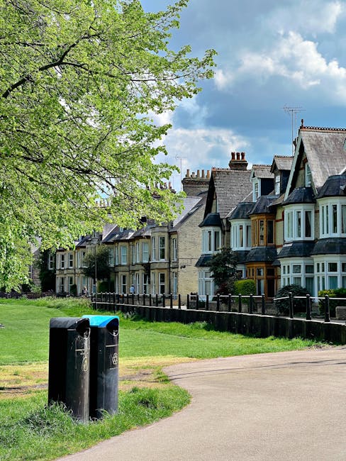 A row of multi-storey terraced houses with bay windows and brick exteriors, situated behind a black metal fence, under a partly cloudy sky. In the foreground, there are two black waste bins with blue lids placed on a grassy area adjacent to a paved footpath. A large leafy tree with green foliage extends over the scene, partially shading the houses and the bins. The image captures a typical residential street scene during daytime, with no visible moving activity or furniture, but it provides context relevant to home relocation and moving services offered by Removal Company Highbury.