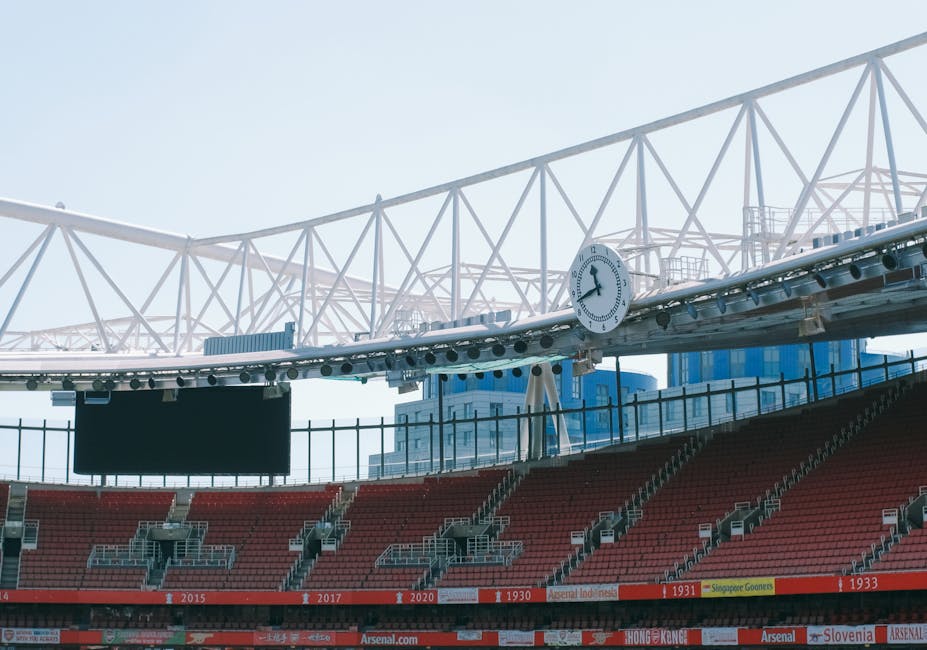 The image shows a section of a stadium with red seating and a partially visible modern white roof structure overhead. In the background, there are tall office buildings made of glass, indicating an urban setting. A large black electronic scoreboard is suspended above the seating area, and a white analog clock mounted on the roof structure displays the time as approximately 1:15. The stadium appears to be prepared for an event, with empty seats and no visible spectators. The structural elements include metal frameworks supporting the roof, with lighting fixtures attached. The overall scene suggests an outdoor sports venue in a city, with the roof designed for weather protection and spectator viewing. This visual could relate to furniture transport or home relocation services, such as those provided by Removal Company Highbury, especially when planning logistics for equipment or event setup.