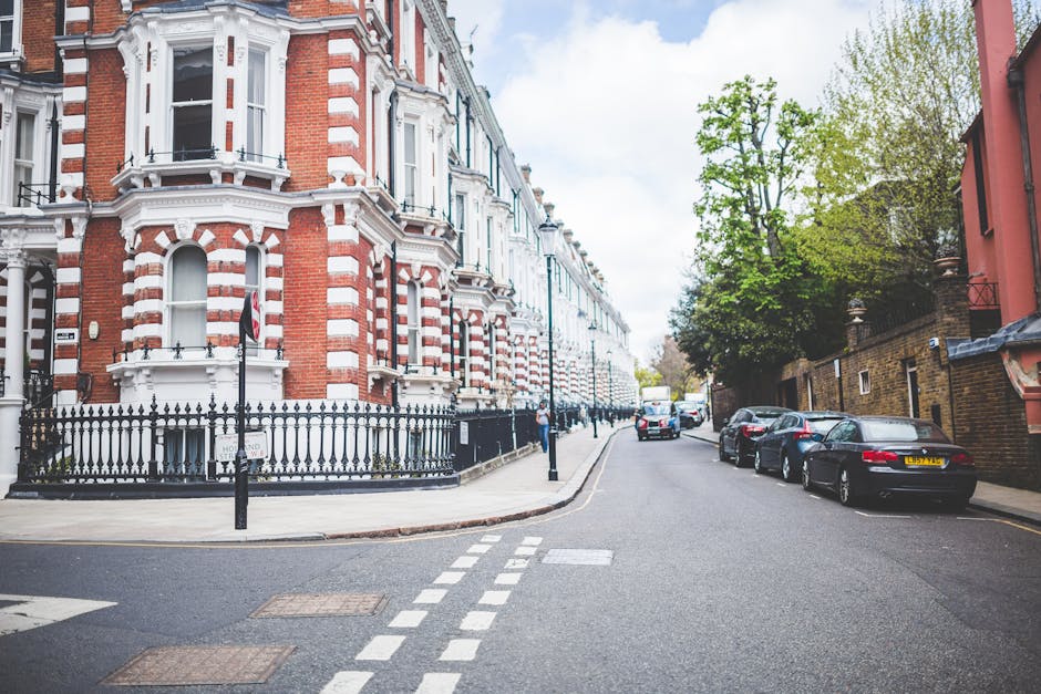 A street view on a narrow Highbury street featuring a row of Victorian-style terraced houses with red brick facades, white decorative trim, and bay windows. In the foreground, a black wrought iron fence encloses the front of a property, with a small sign indicating 'houser cord.' The street has parked cars along the right side, including black, silver, and grey vehicles, lining the curb. The pavement is clean, with a pedestrian crossing marked by white dashed lines approaching the intersection. On the left, a person is seen walking on the sidewalk adjacent to the property, which is part of a house removal or house relocation process. The scene is illuminated by natural daylight, with a partly cloudy sky overhead and trees with green foliage lining the street, providing a calm residential environment. This setting showcases typical urban household street access, relevant to the services of Removal Company Highbury specializing in removals and furniture transport through narrow street environments.